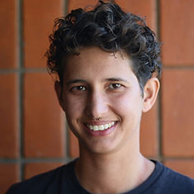 Cássio Kelm, jovem de cabelos castanhos escuros e ondulados, sorrindo, com fundo de parede em tom alaranjado.