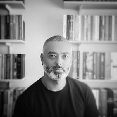 Henrique dos Santos, homem de cabelos grisalhos, com barba e cabeça raspada, em retrato preto e branco com estante de livros ao fundo.
