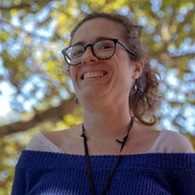 Jade Azevedo, mulher de cabelos castanhos, usando óculos, sorrindo ao ar livre, com árvores desfocadas ao fundo.