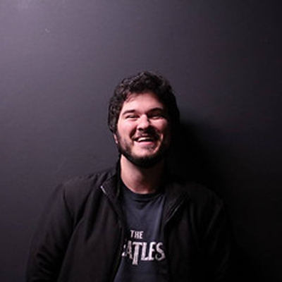 William Biagioli, homem jovem de cabelos escuros e barba curta, sorrindo em retrato com fundo escuro.